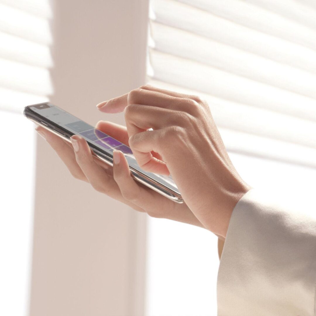 A person using their cell phone in front of blinds.